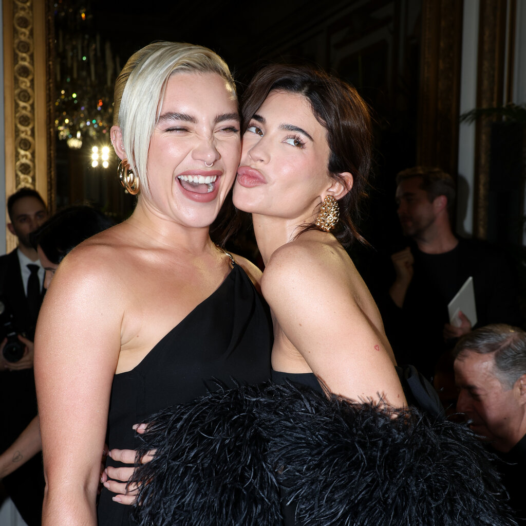 Florence Pugh in off-the-shoulder black dress and Kylie Jenner in a feathered gown hug each other and pose for the camera. 