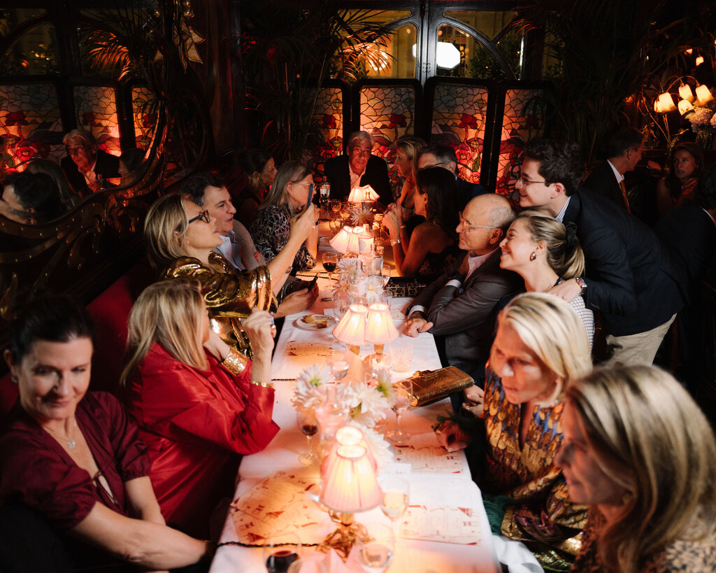 A group of well-dressed people seated at a long dinner table inside a dimly lit restaurant. 