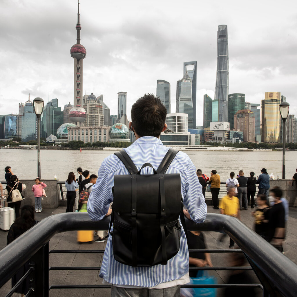 The a view from the back of a man with short hair, wearing a striped shirt and a black backpack, ahead of him is the Shanghai skyline. 
