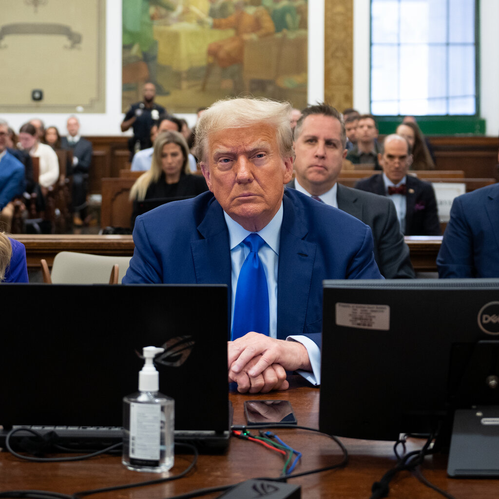 Donald J. Trump, wearing a blue suit and tie, sits at a courtroom table between two lawyers as other people sit in the gallery behind them.
