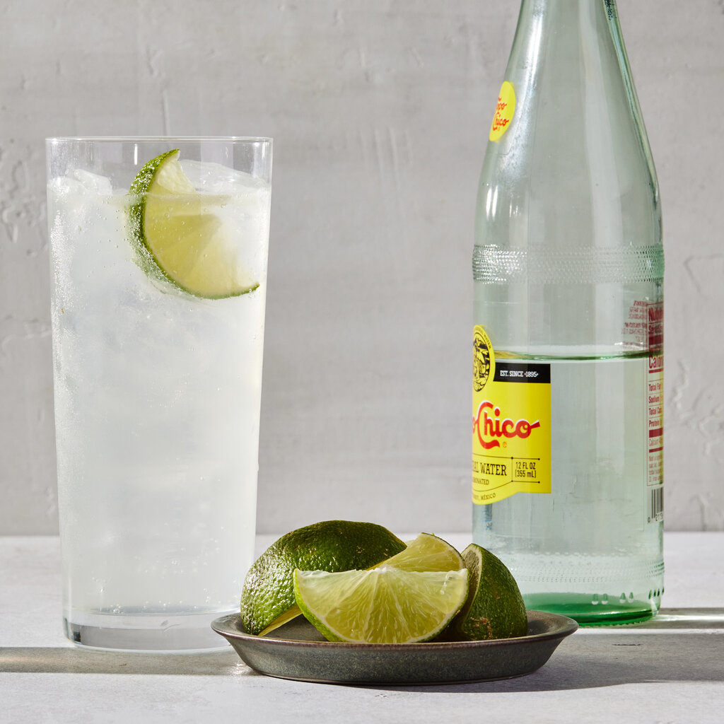 A glass and a bottle on white background. In the foreground a plate of sliced limes.