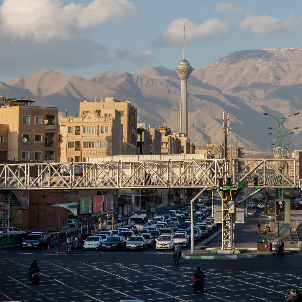 A landscape image of Tehran. Cars wait at a stop lights and in the background large buildings are dwarfed by a mountain range. 