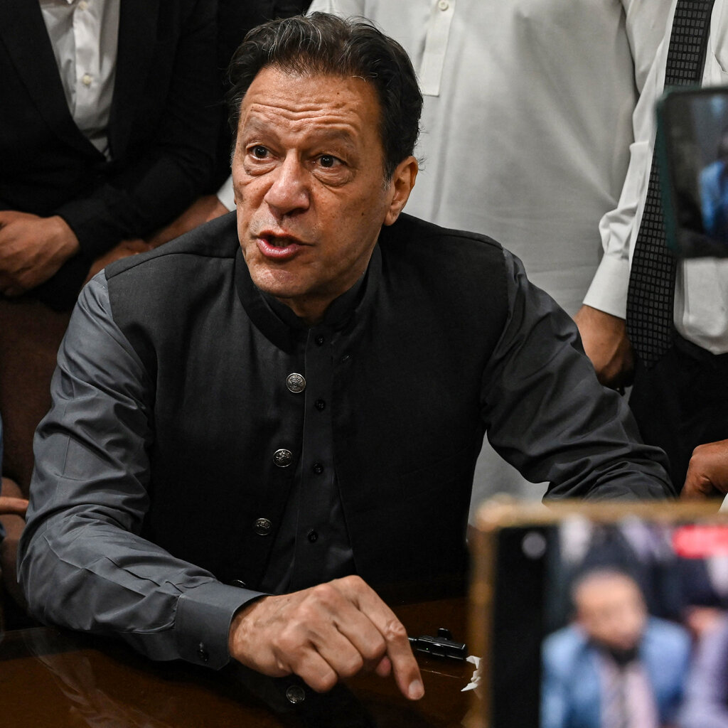 Imran Khan, dressed in black, sits among a group of men who are standing, as a couple of them film him on their cellphones.