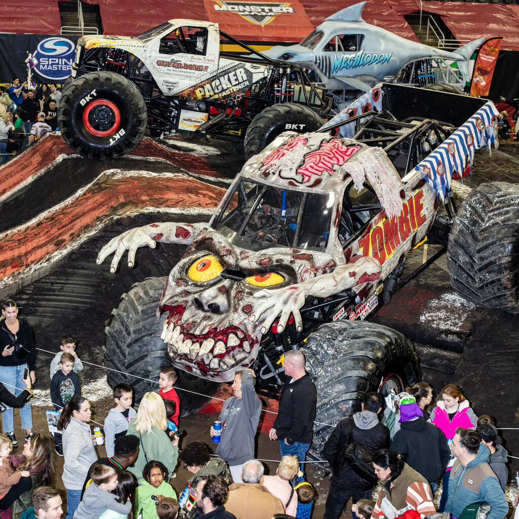 A small crowd gathered in a penned-off area beside three monster trucks, one decorated to look like a zombie, with yellow eyes and arms extending from its body.