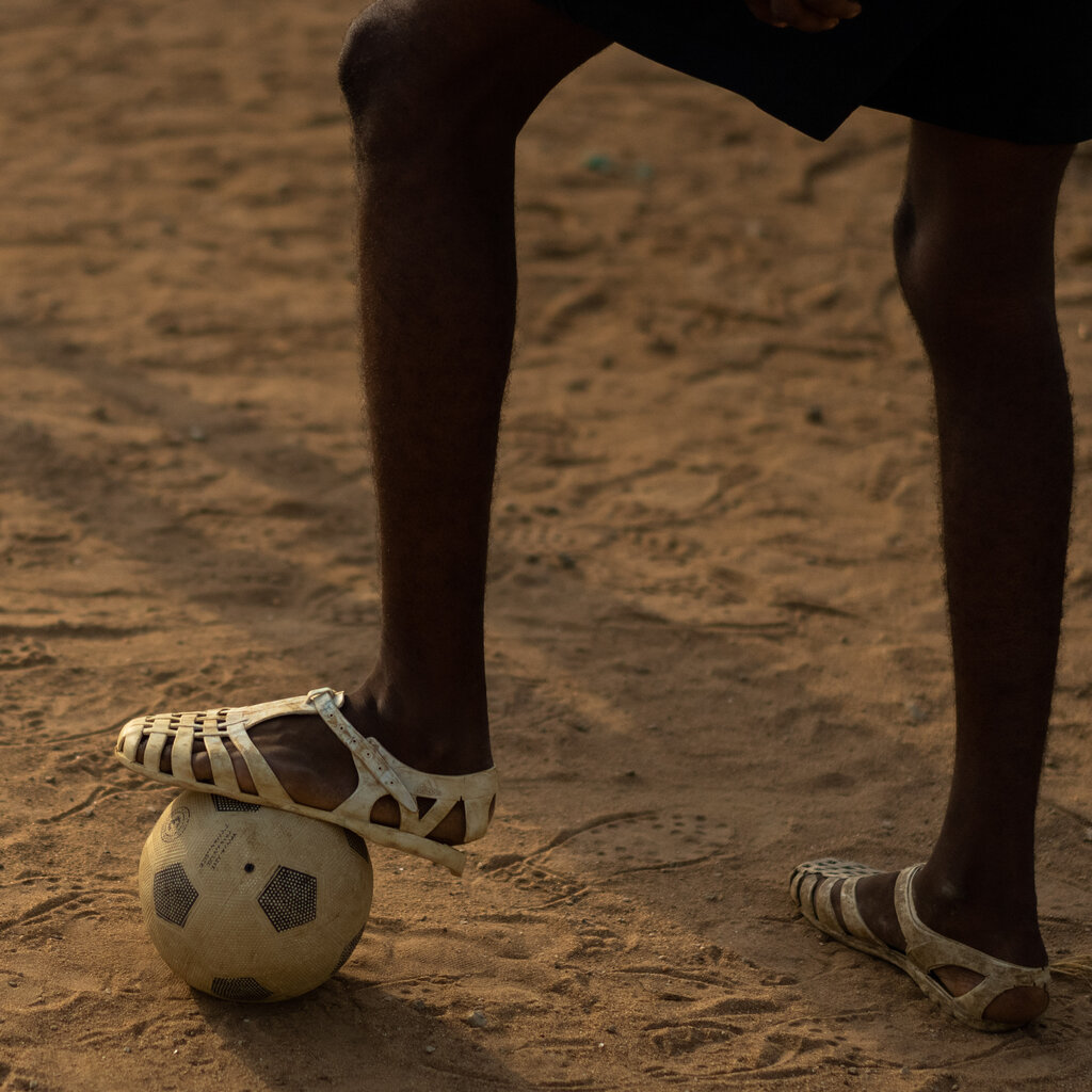 A soccer player stands over a dirty soccer ball on a dusty field covered in footprints. He is wearing white plastic sandals known as lêkê.