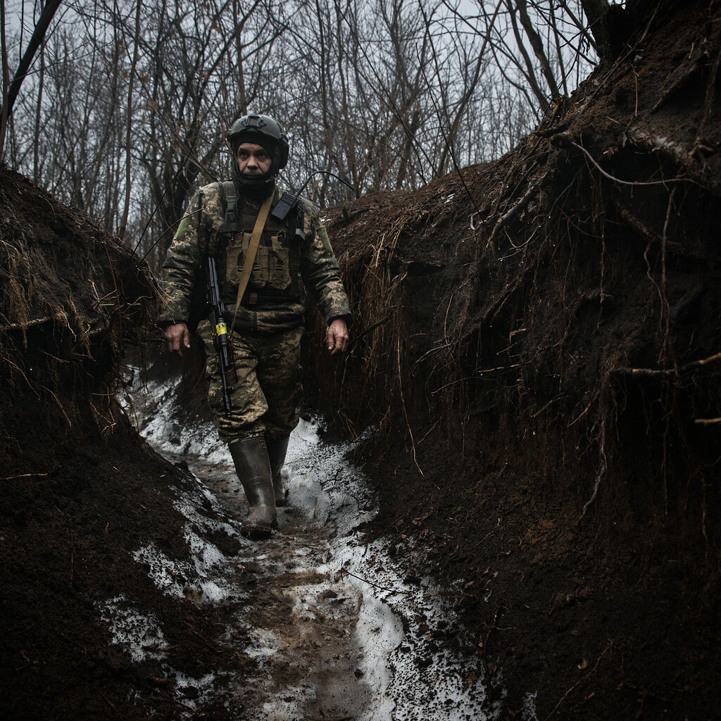 A Ukrainian soldier in camouflage walks through a shoulder-deep trench in a barren forest.