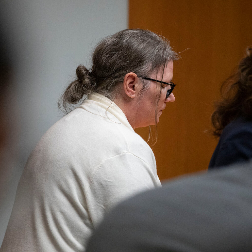 The side profile of a woman with glasses and long hair in a low bun looking down in a courtroom.