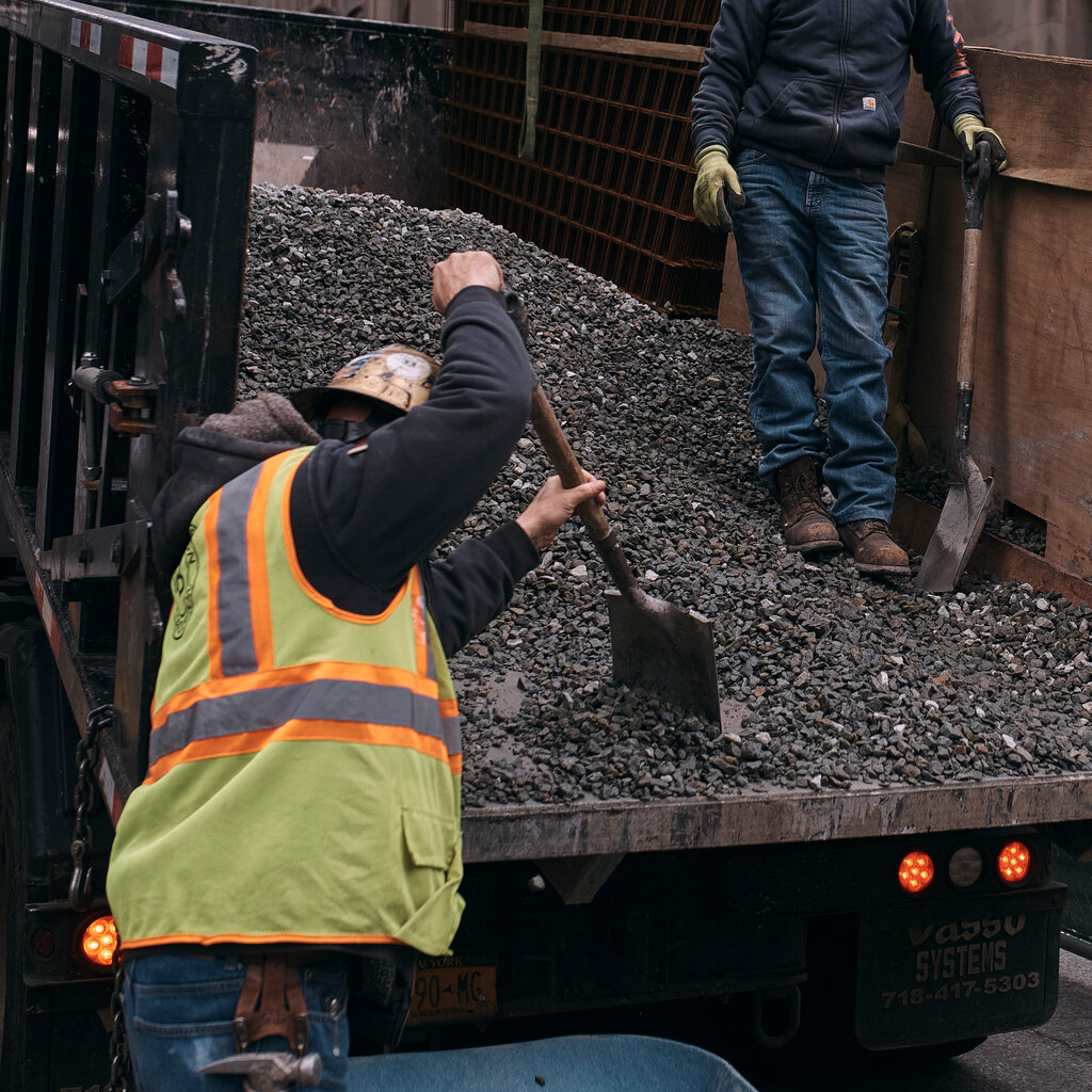 Two men in hard hats shovel gravel from the back of a dump truck.