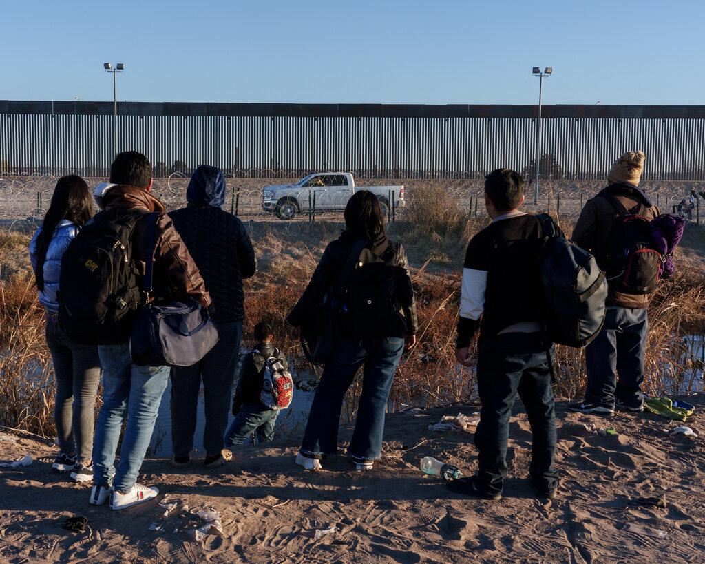 A group of people standing at the foot of a river look at a truck parked in front of a border wall.