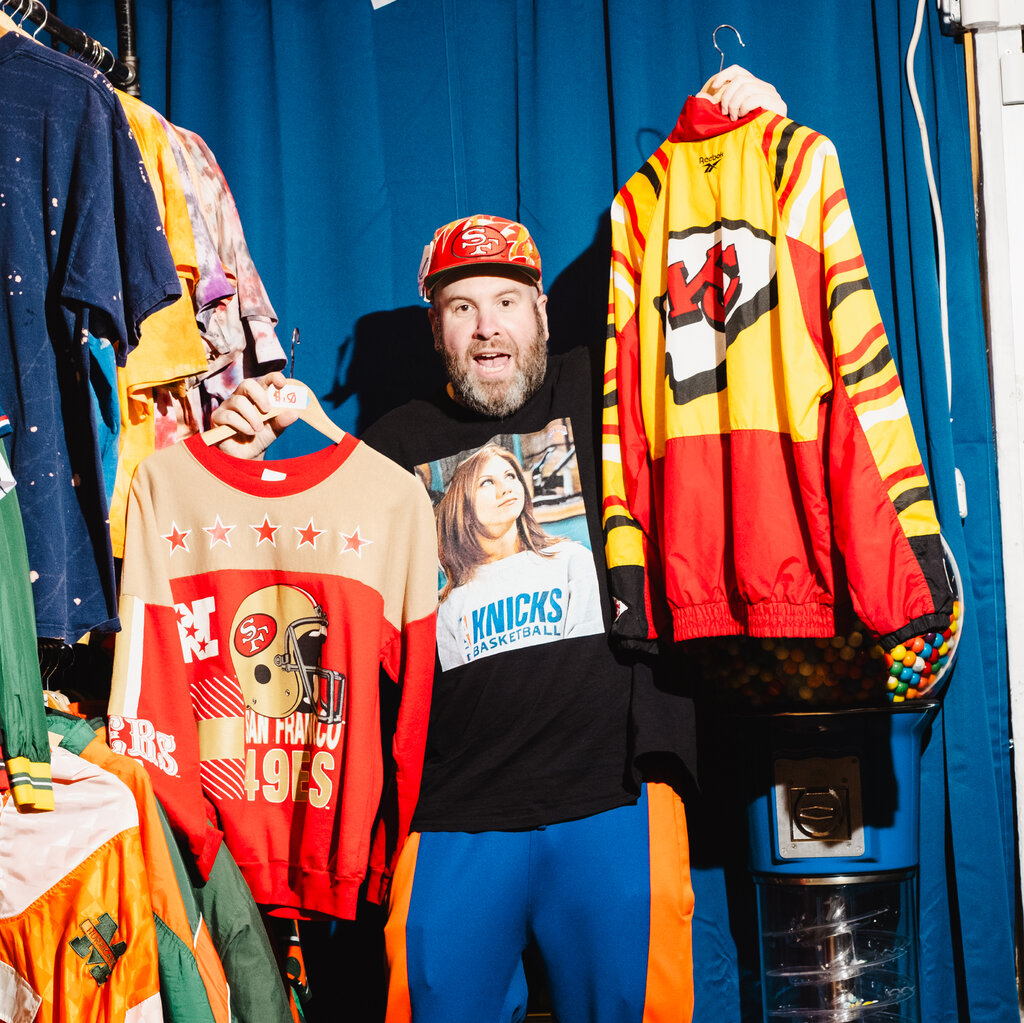 A bearded man decked out in various team sportswear holds a long-sleeve red and beige 49ers T-shirt in one hand and a red and yellow Chiefs jacket in the other. Next to him are racks of colorful sportswear.