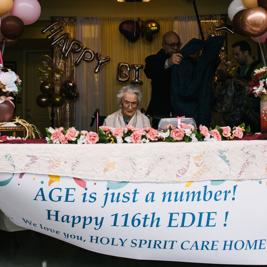 Edith Ceccarelli sits behind a table covered in roses and surrounded by balloons. A banner on the front of the table reads: “AGE is just a number! Happy 116th EDIE! We love you, HOLY SPIRIT CARE HOME”