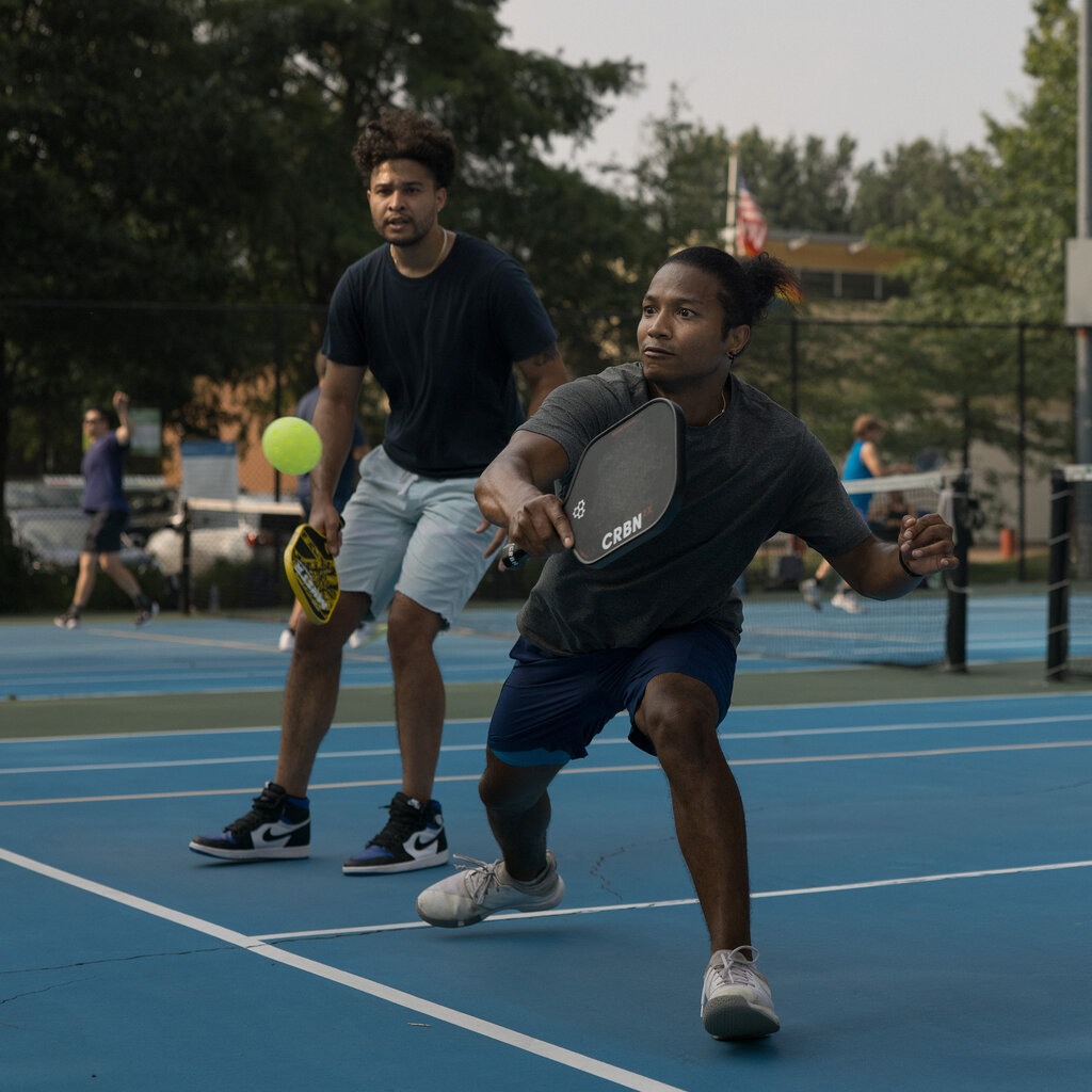 Two players wearing shorts play pickleball on a blue court. One lunges for the ball.