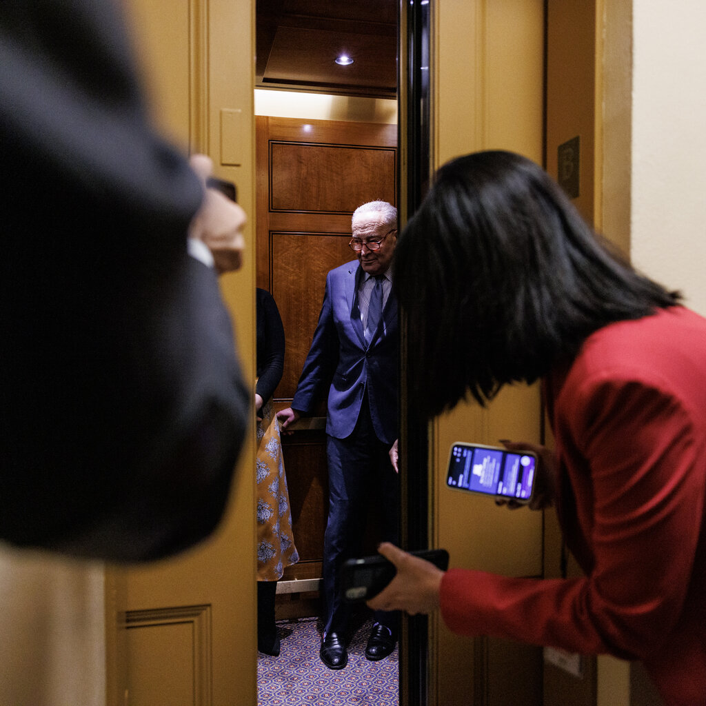 Chuck Schumer stands in an elevator, obscured by the doors. A woman wearing a red blazer peers in. 