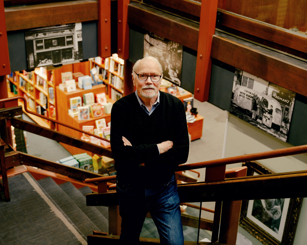 A man with short white hair and beard, wearing a dark sweater, poses on a staircase in the middle of a bookstore. Behind him are shelves with books.