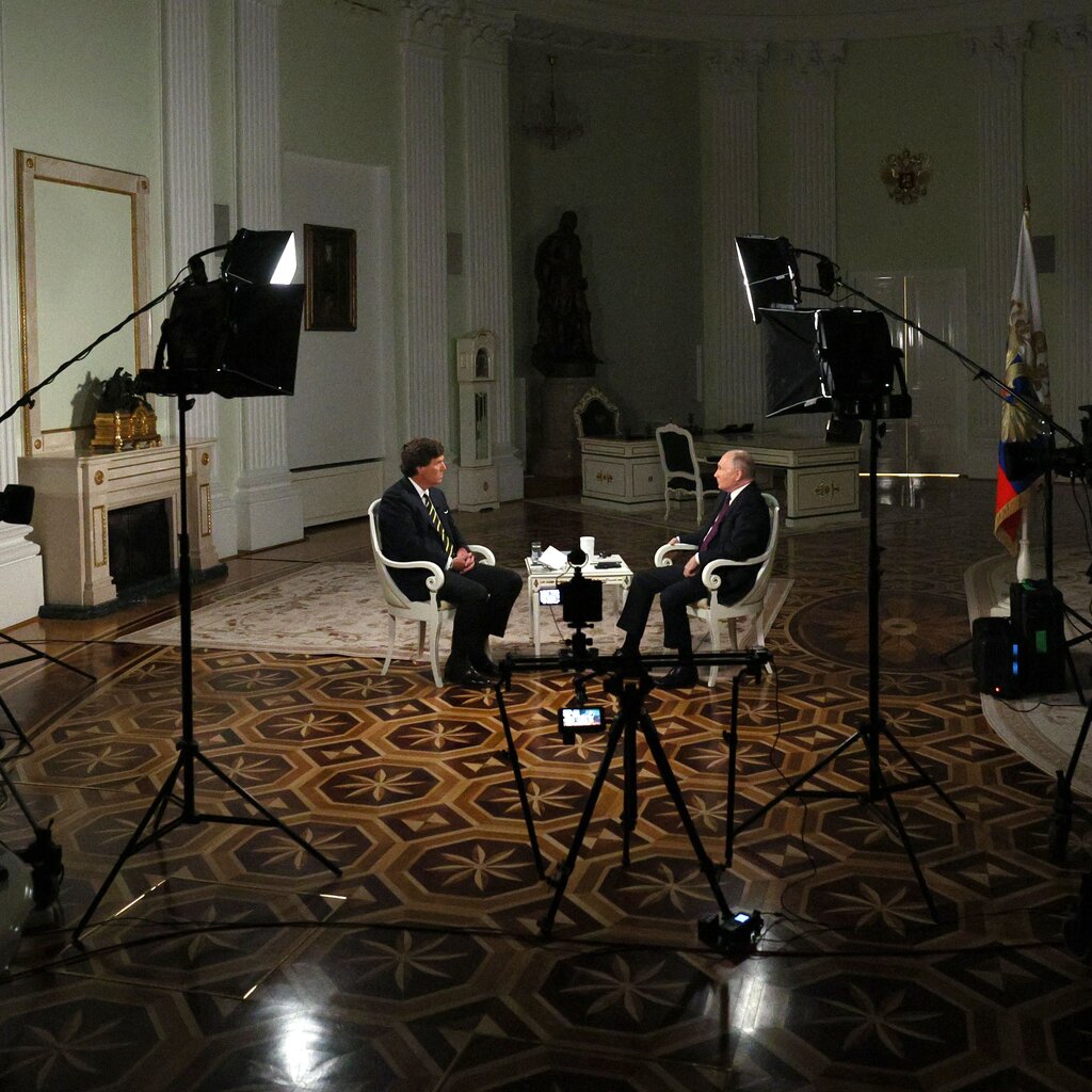 Tucker Carlson and Vladimir Putin in suits sitting across from each other. 