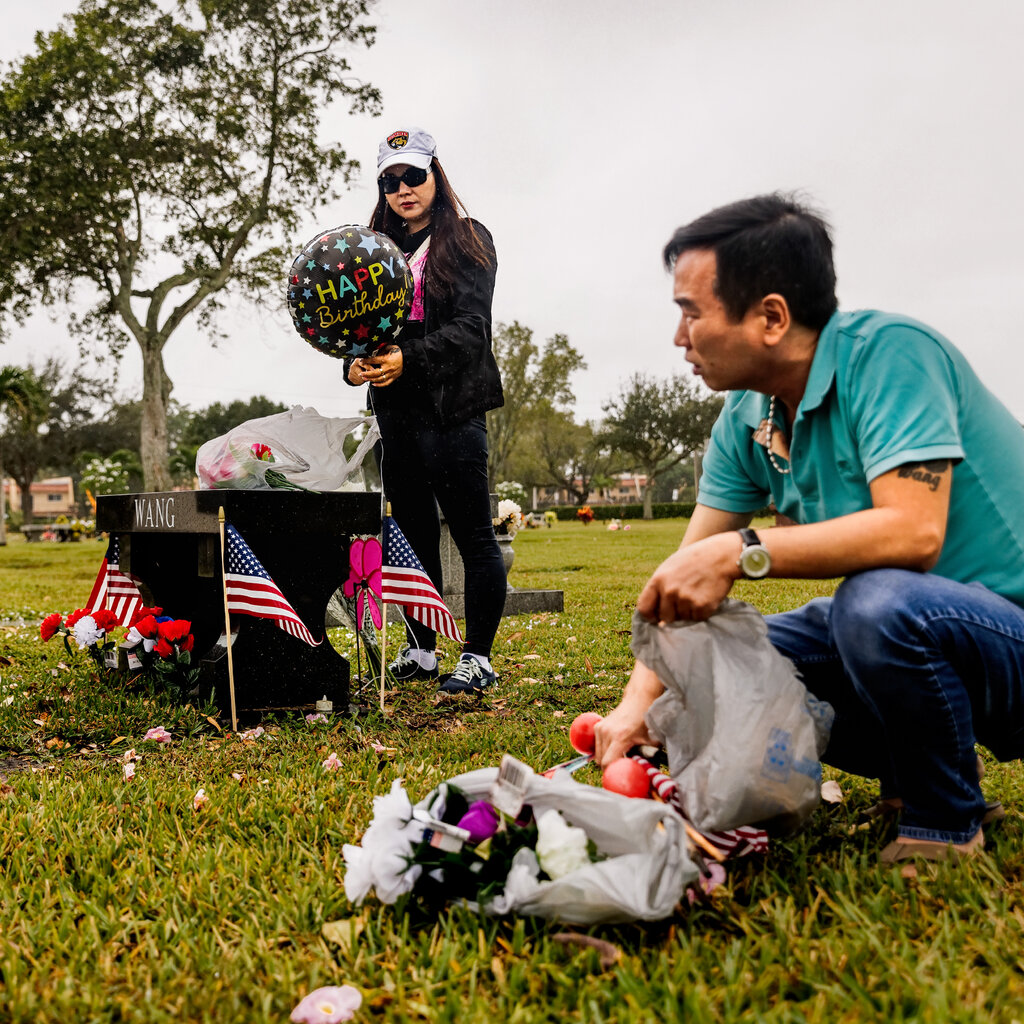 A woman holding a birthday balloon stands by a black memorial table in a graveyard. Nearby, a man crouches over a bag with flowers and other items. 