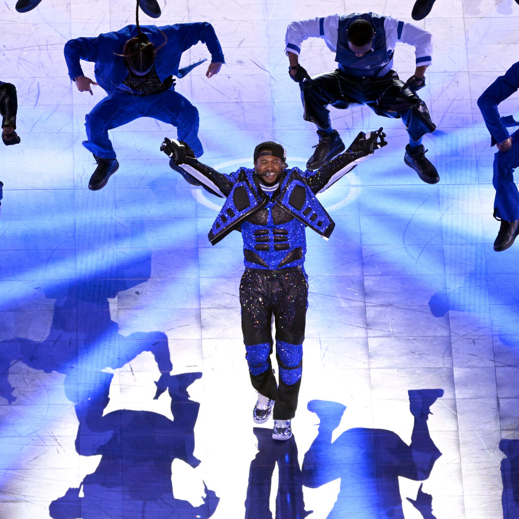 A man dressed in a blue-and-black motorcycle outfit stands with his arms out as dancers surround him on a stage.