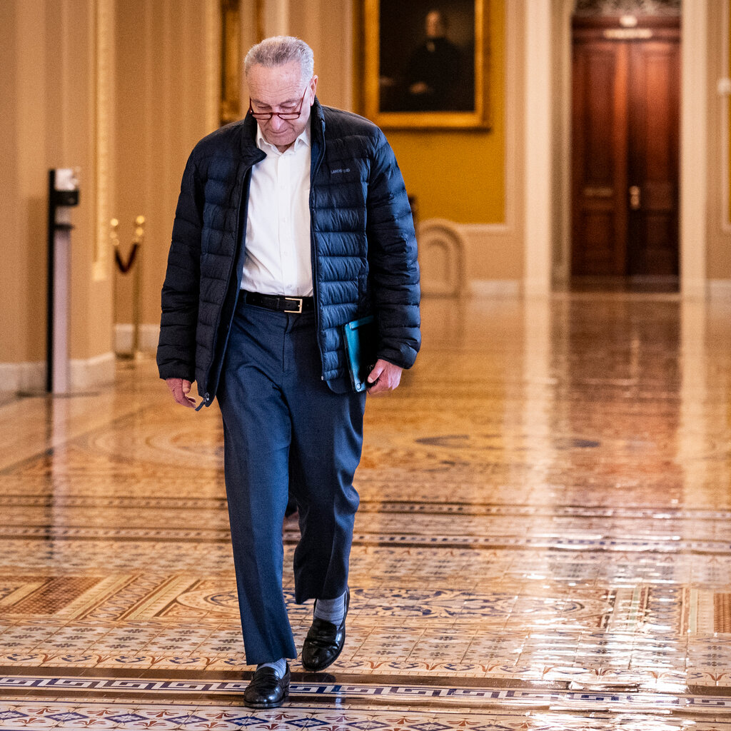 Chuck Schumer, looking down, walks through a corridor with a tiled floor in the Capitol.