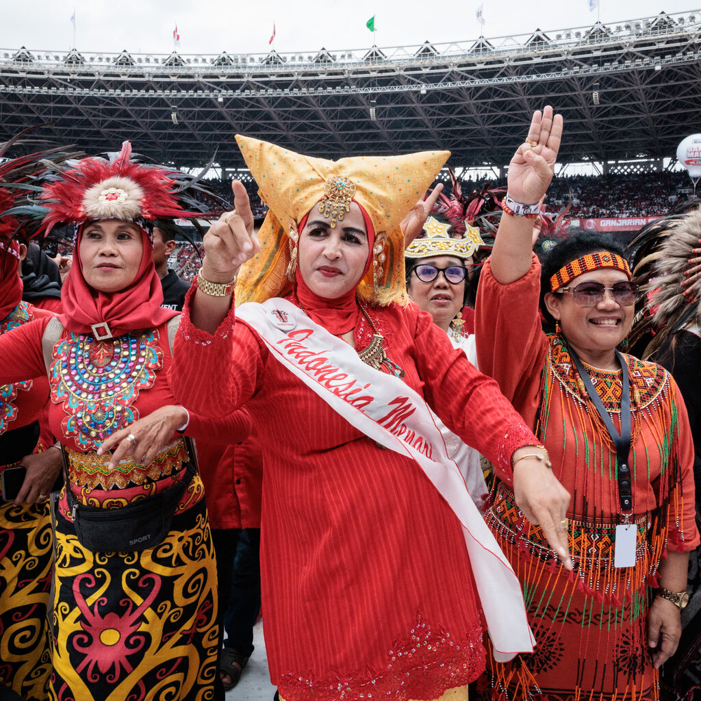 Many people in a stadium. A group closest to the camera wears colorful costumes and fanciful headdresses, several with feathers.