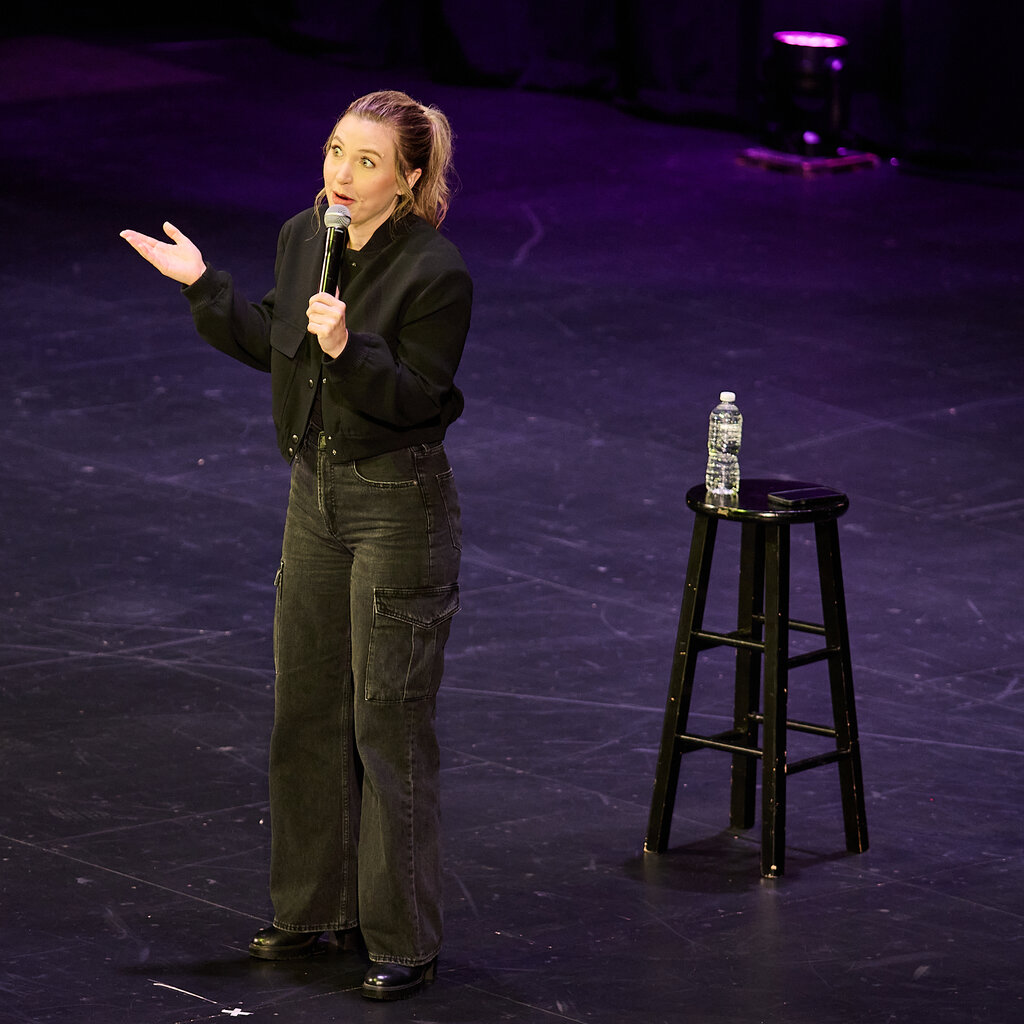 The comedian Taylor Tomlinson speaks into a microphone with one arm raised in a shrug onstage. In the background, a single stool with a bottle of water placed on the seat.