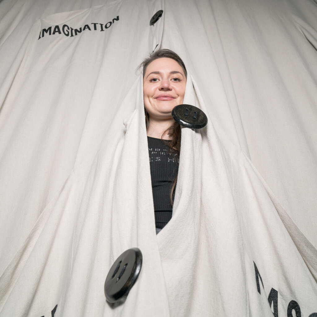 A brunette woman peeks her head out from inside a gargantuan white button-down shirt that reads “Imagination” on the front pocket.