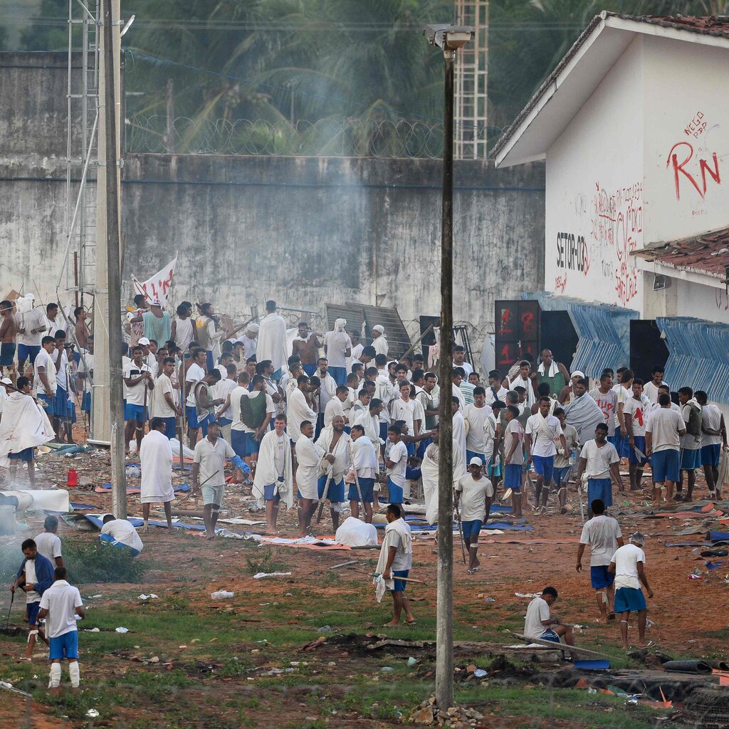A crowd of men in white shirts and blue shorts stand in a yard bordered by a tall cement wall topped with barb-wire in places. 