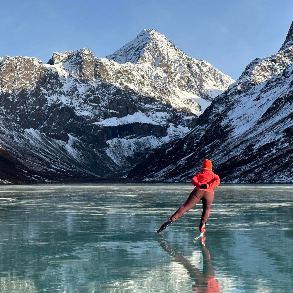 A lone woman in a red jacket skates across the ice of a frozen lake. Ahead of her rugged, snowy mountains rise into the clear sky.