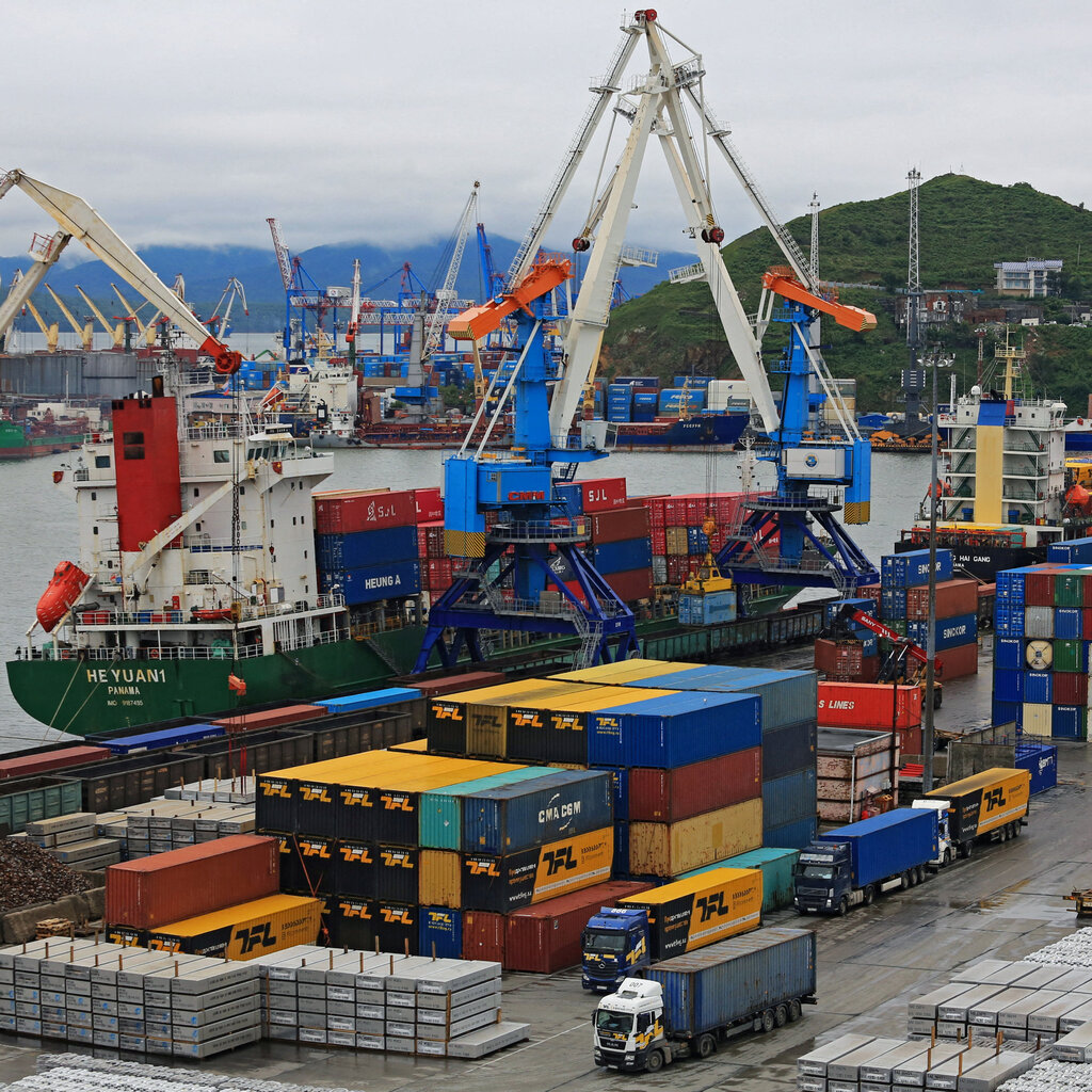 A shipping port with containers stacked high. In the background, multiple ships and cranes. 