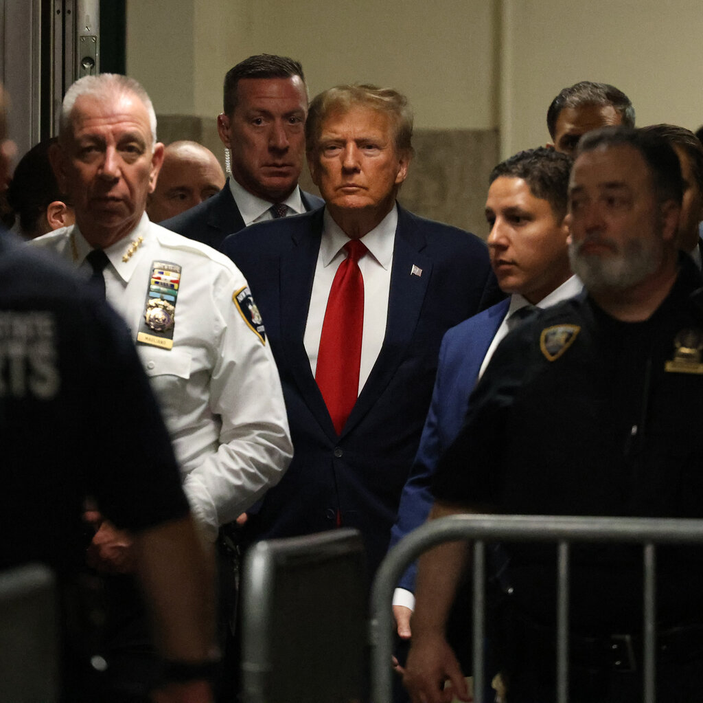 Donald Trump, in a dark suit and red tie, with his attorneys, escorted by police officers.