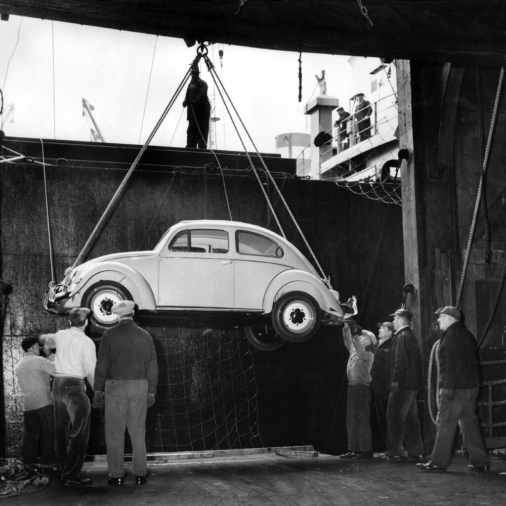 A Volkswagen Beetle is lowered from a ship at a port in a black-and-white photo.
