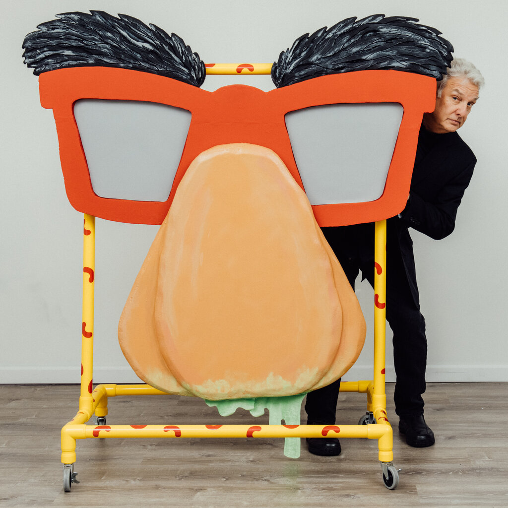 A man dressed in black is peeking out from the side of one of the scenic elements of his new show. It’s a giant nose, topped with red-frame eyeglasses and bushy black eyebrows. Green slime is seemingly dripping from the nose’s nostrils.