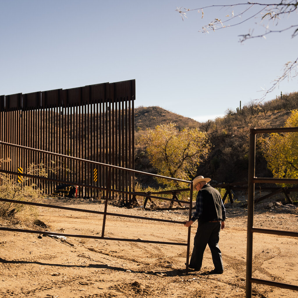 Jim Chilton, in a cowboy hat, opening a large metal gate. Behind him is the border wall.