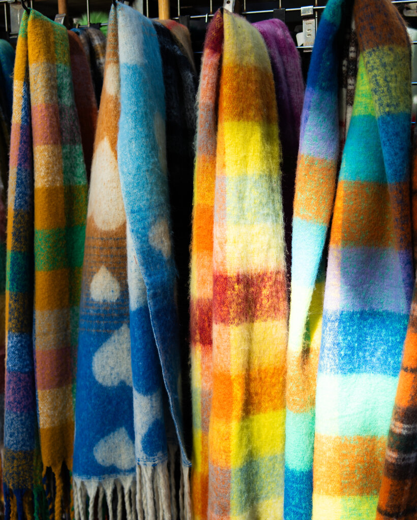 Several versions of fuzzy rainbow-check scarves hanging on a street display rack.