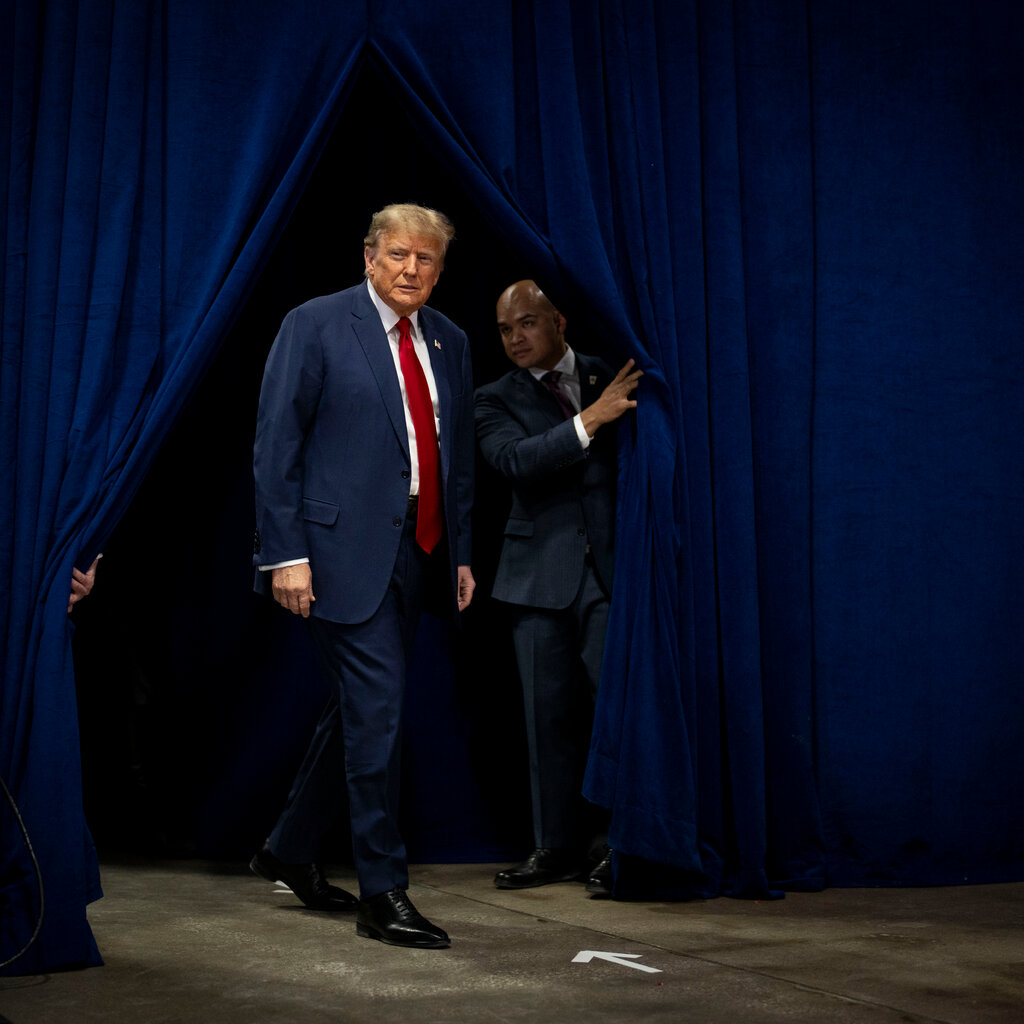 Donald Trump in a suit and tie walks out from a curtain being held open by two people. 