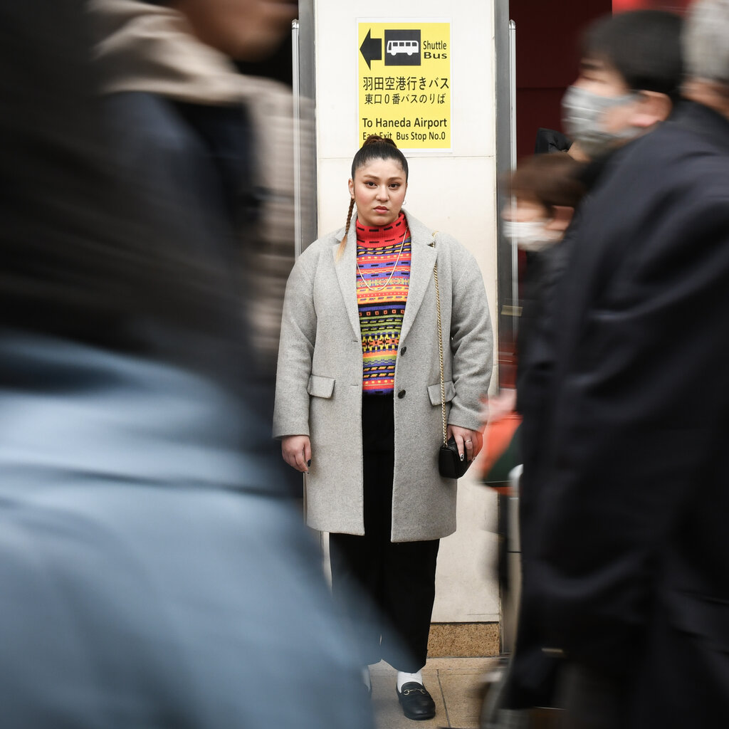 A woman in a gray coat stands still as people in masks walk past her. 