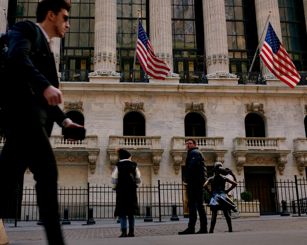 American flags wave in the wind on the front of the New York Stock Exchange while pedestrians walk by. 