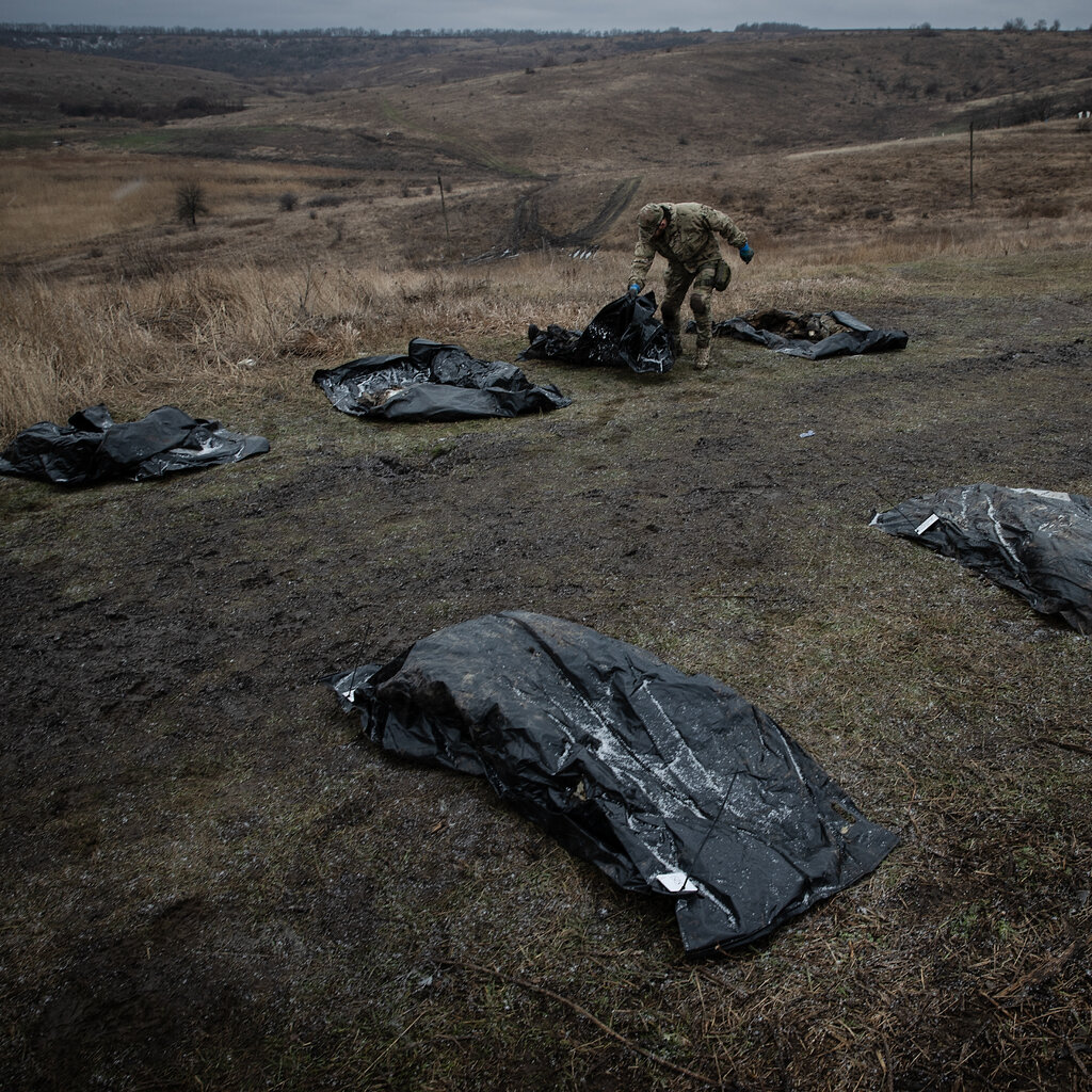 A person in fatigues collects dead bodies covered in black plastic in a rolling field of yellowing scrub.