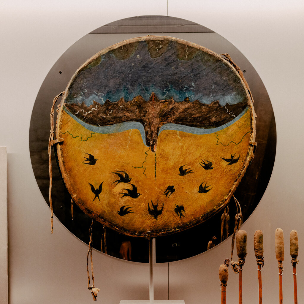 A display shows a large round drum with a large bird with brown feathers facing down across its top half, against a blue background, and silhouettes of a dozen small birds against a yellow background across the bottom. Five padded drumsticks sit below it. 