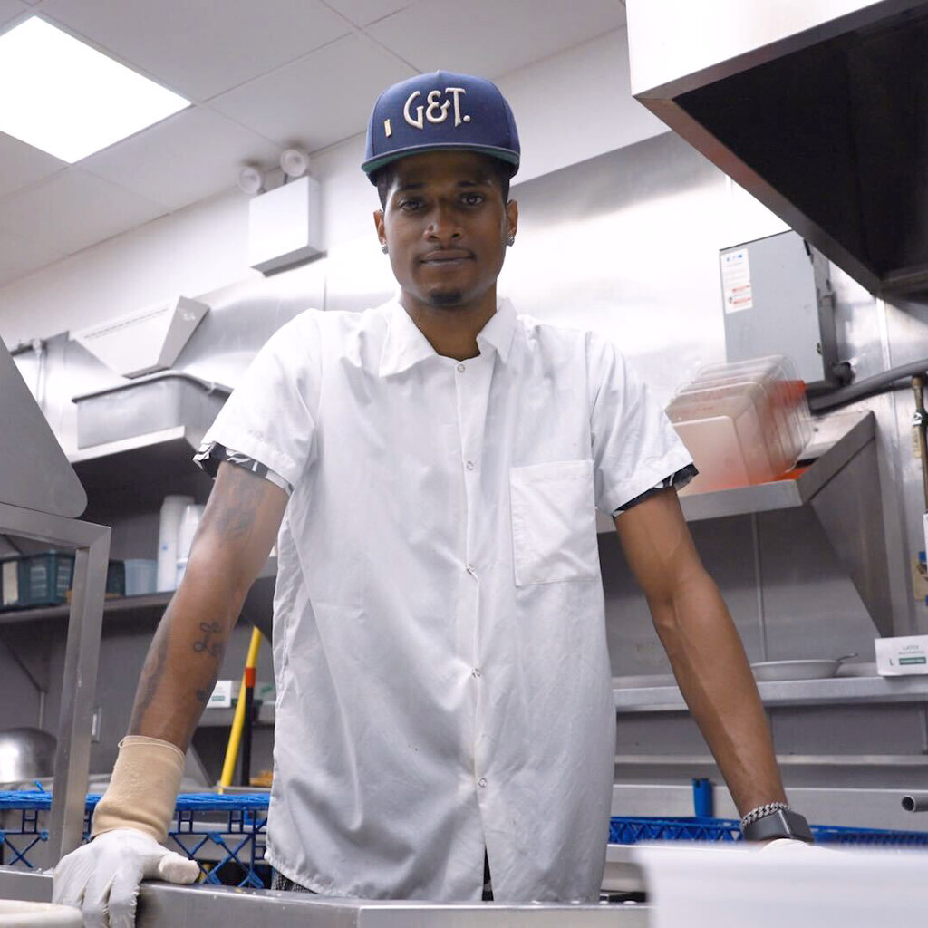 Drevon Alston, a tall Black man in a white button-down work shirt and a blue oversize baseball hat that says “G&T,” stands behind the dishwashing station in a commercial kitchen.