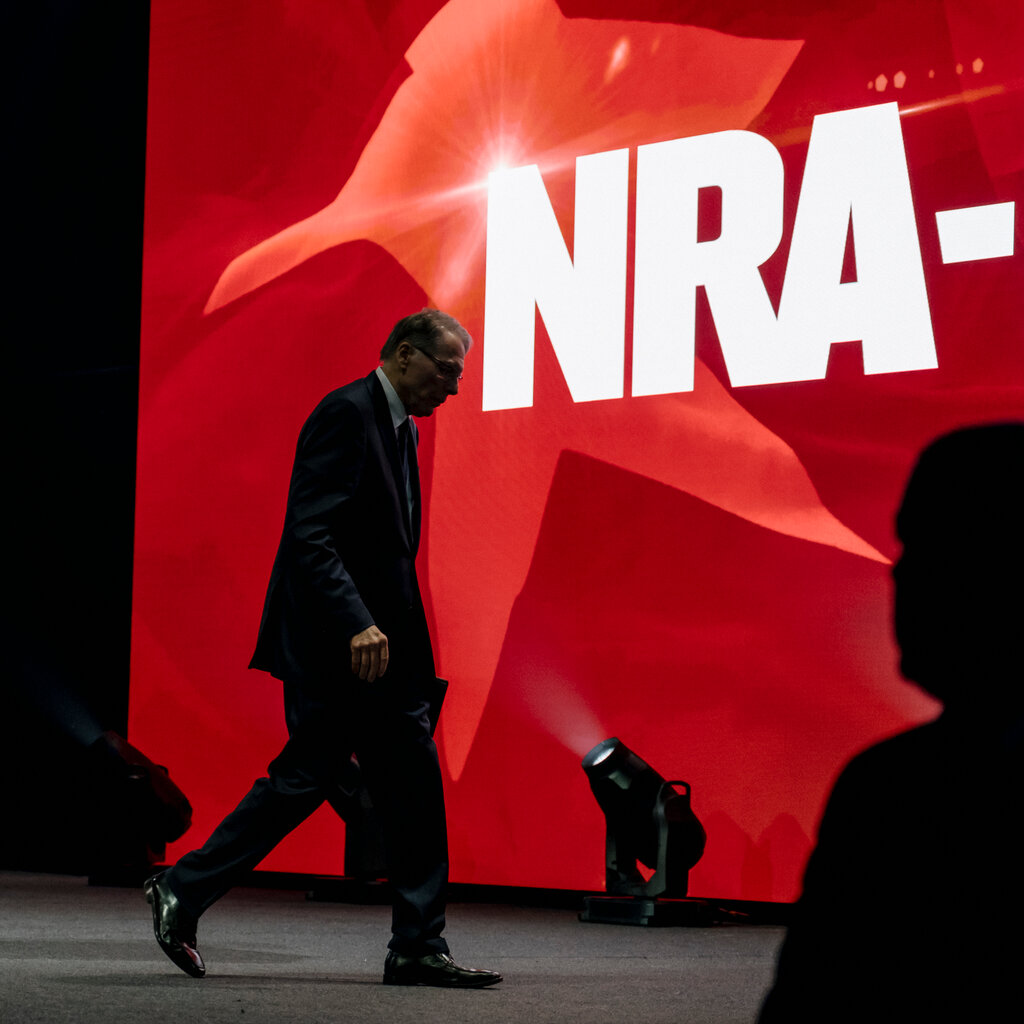 A man in a suit and glasses walks across a stage. A large screen behind him reads “N.R.A.”
