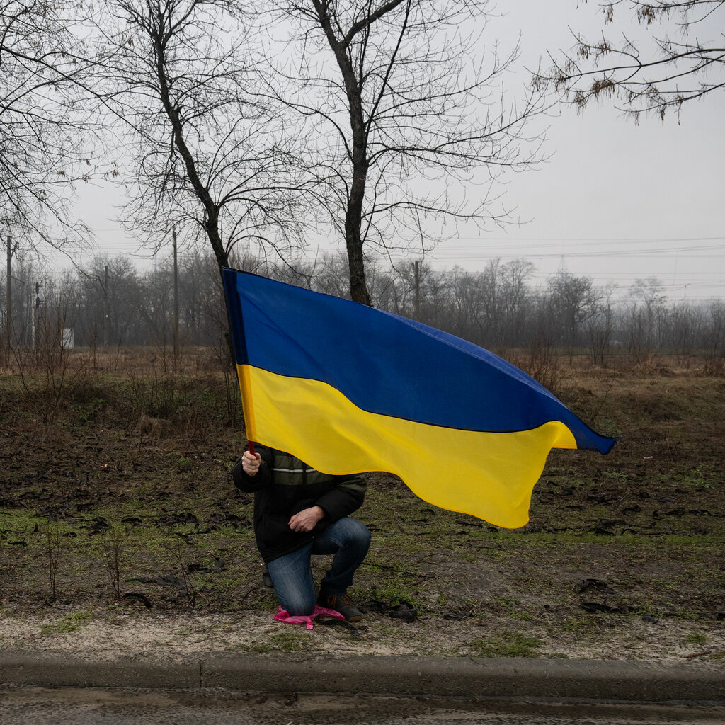 A person carrying a blue and yellow Ukrainian flag kneels by the side of a road. 