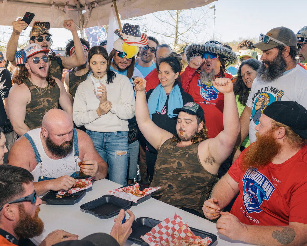 Several contestants are seated around a table with boxes of pork. One man in the center has his arms in celebration. Several others are behind the contestants, cheering them on.