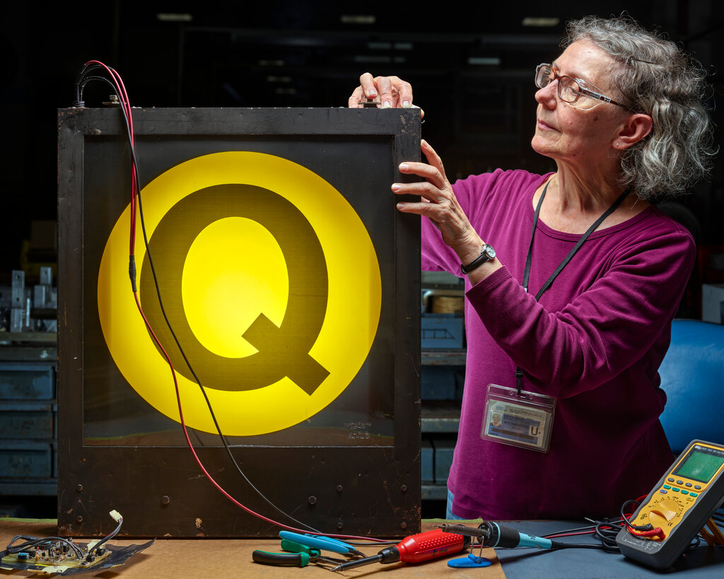 A woman in safety glasses and a magenta shirt works on a box with an illuminated yellow “Q” subway sign.