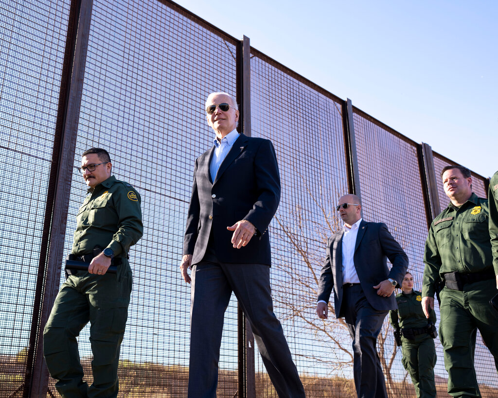 President Biden surrounded by border patrol agents walking near a border fence.