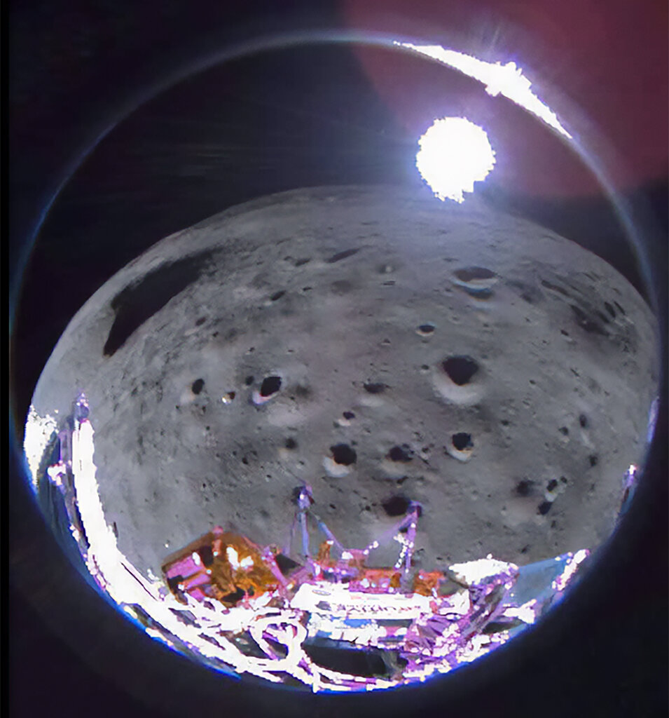 A fish-eye view from an onboard camera of the Intuitive Machines Odysseus lander, with the curved, pockmarked surface of the moon below and a bright spot of light on the horizon.