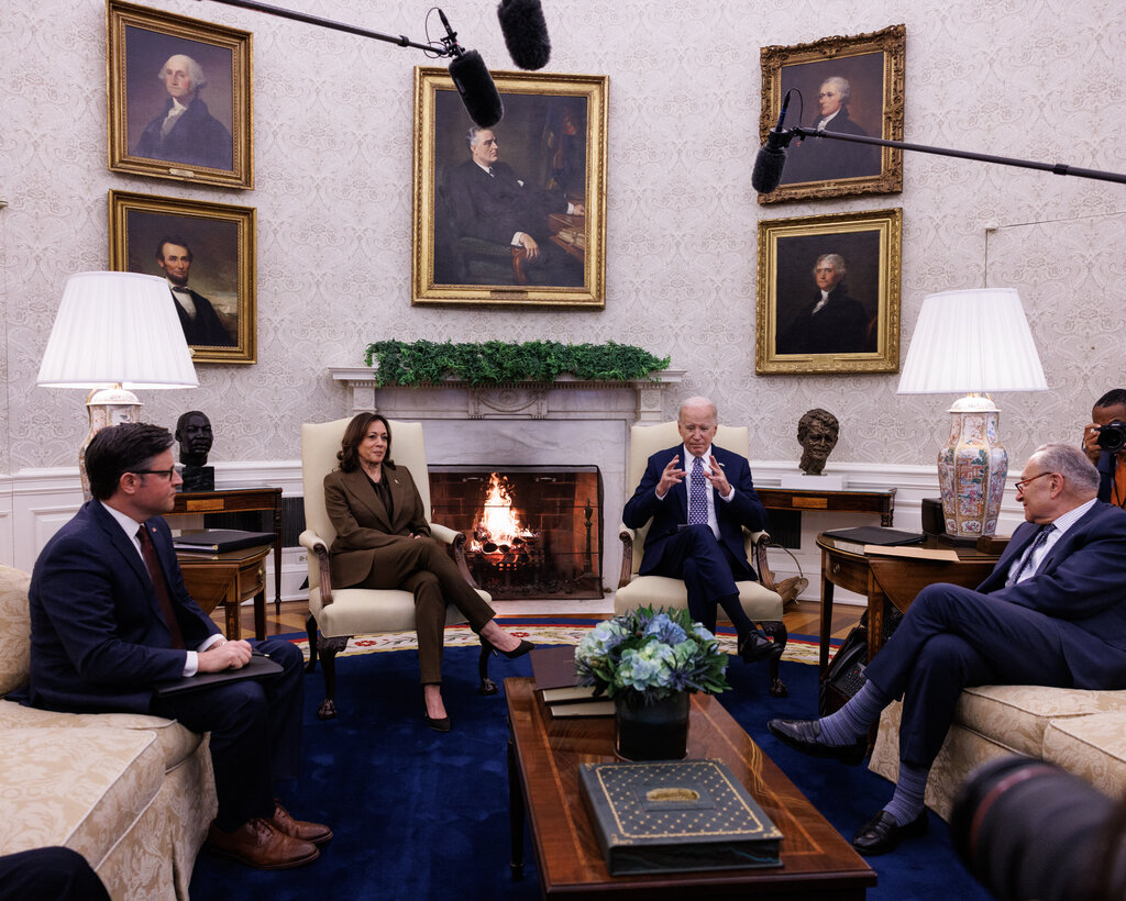 House Speaker Mike Johnson, Vice President Kamala Harris, President Biden and Senator Chuck Schumer sitting around a coffee table in the Oval Office, with a fire in the fireplace at the center.