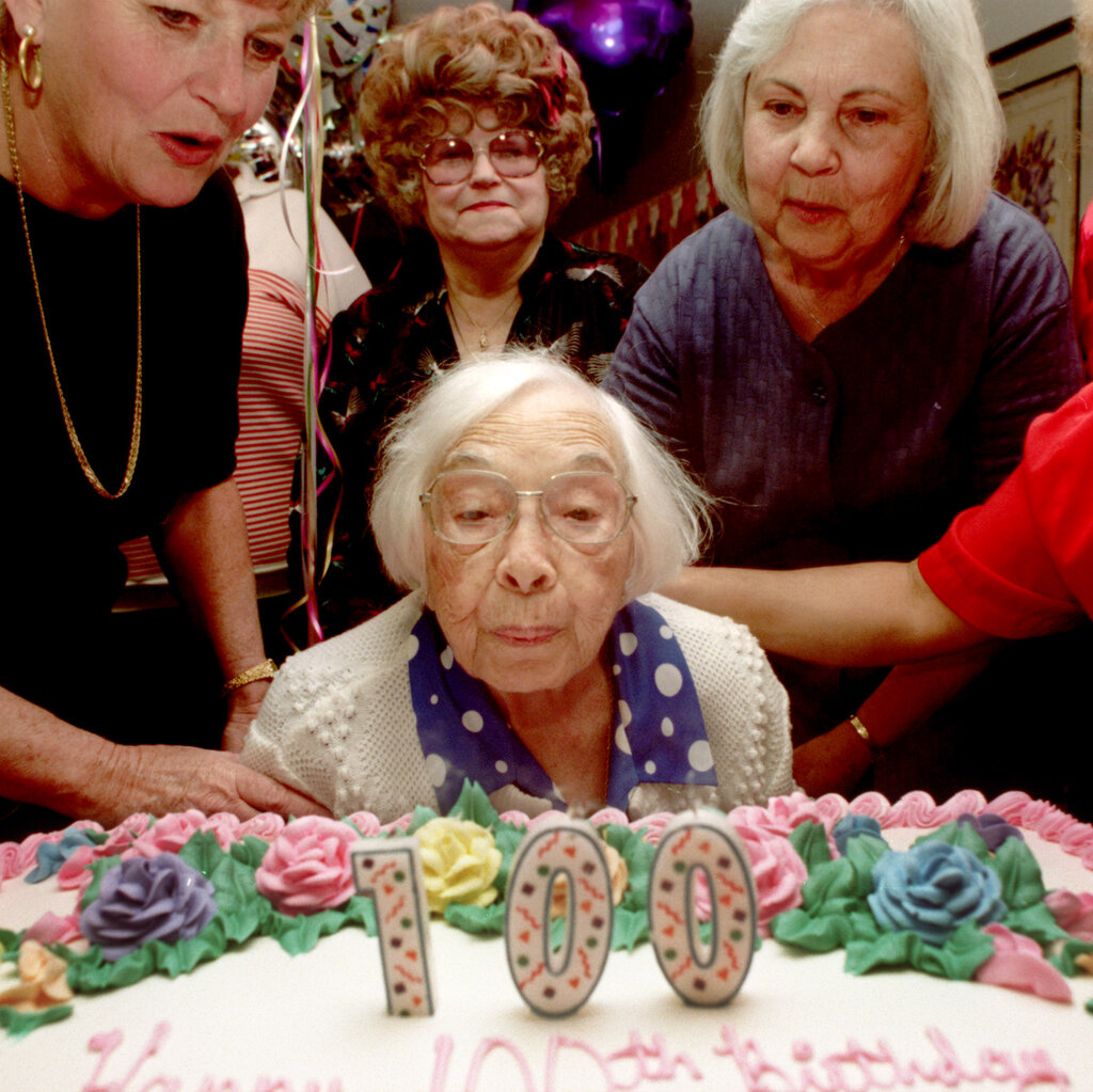 An older woman blows out candles spelling out the number “100” on a birthday cake, surrounded by well-wishers.