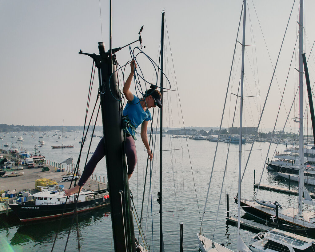 Cole Brauer, in a blue shirt and dark pants, hangs near the top of the mast of a boat in a marina.
