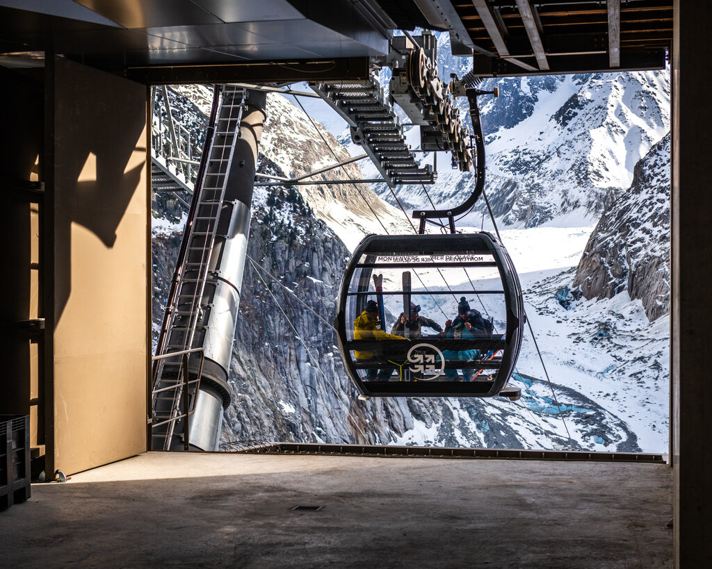A view of a gondola as it leaves a station and descends into a snowy valley surrounded by rugged mountains.