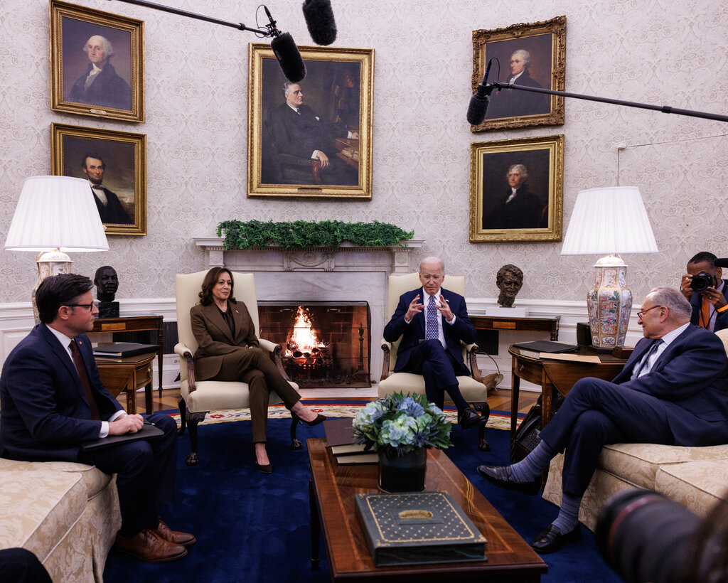 President Joe Biden and Vice President Kamala Harris meeting with Speaker Mike Johnson and Senator Chuck Schumer at the White House. A cluster of microphones is over their heads and several gold-framed portraits of past presidents are on the wall. A fireplace is in the background.